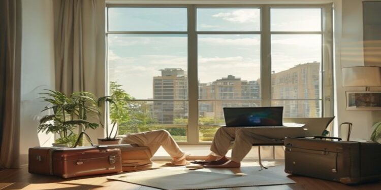 Remote worker using a laptop beside a window with luggage and travel notebook nearby